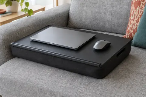 Grand plateau de genoux noir en simili cuir fixé sur un épais coussin, avec un ordinateur portable fermé et une souris, posé sur un canapé gris clair