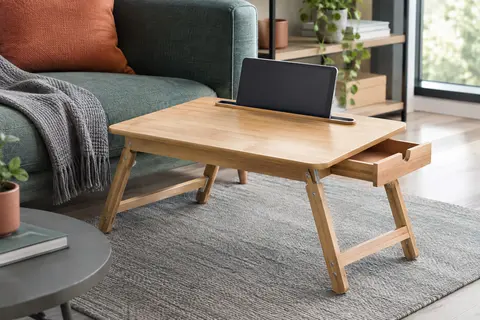 Table de lit en bois à pieds pliants, avec rainure pour tablette et tiroir intégré, près d’un canapé.